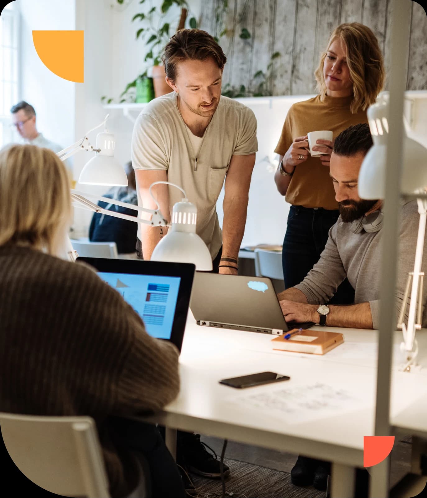Four people collaborate around a table with laptops and papers in a modern, brightly lit office space.