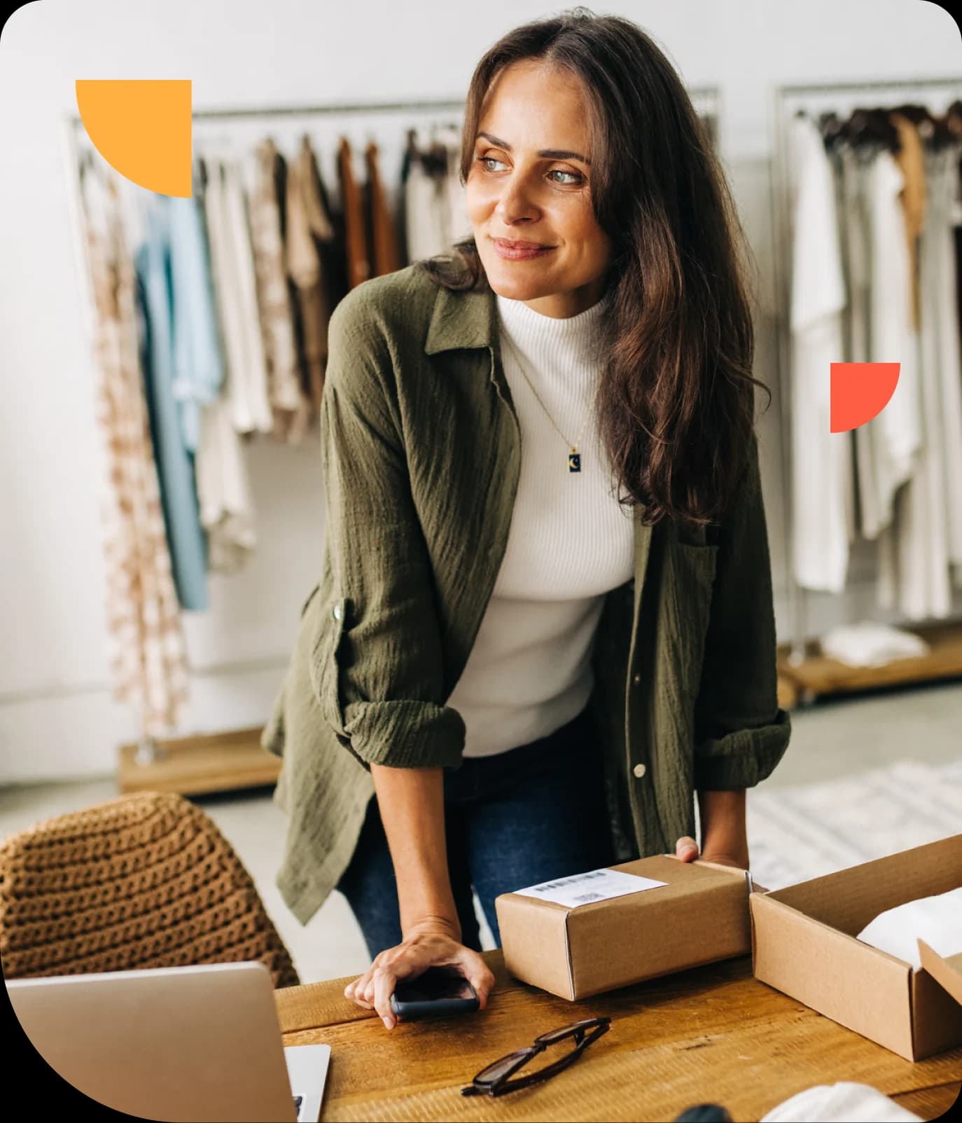 Woman in a green shirt leans on a table with packages, smiling. Clothes hang in the background.