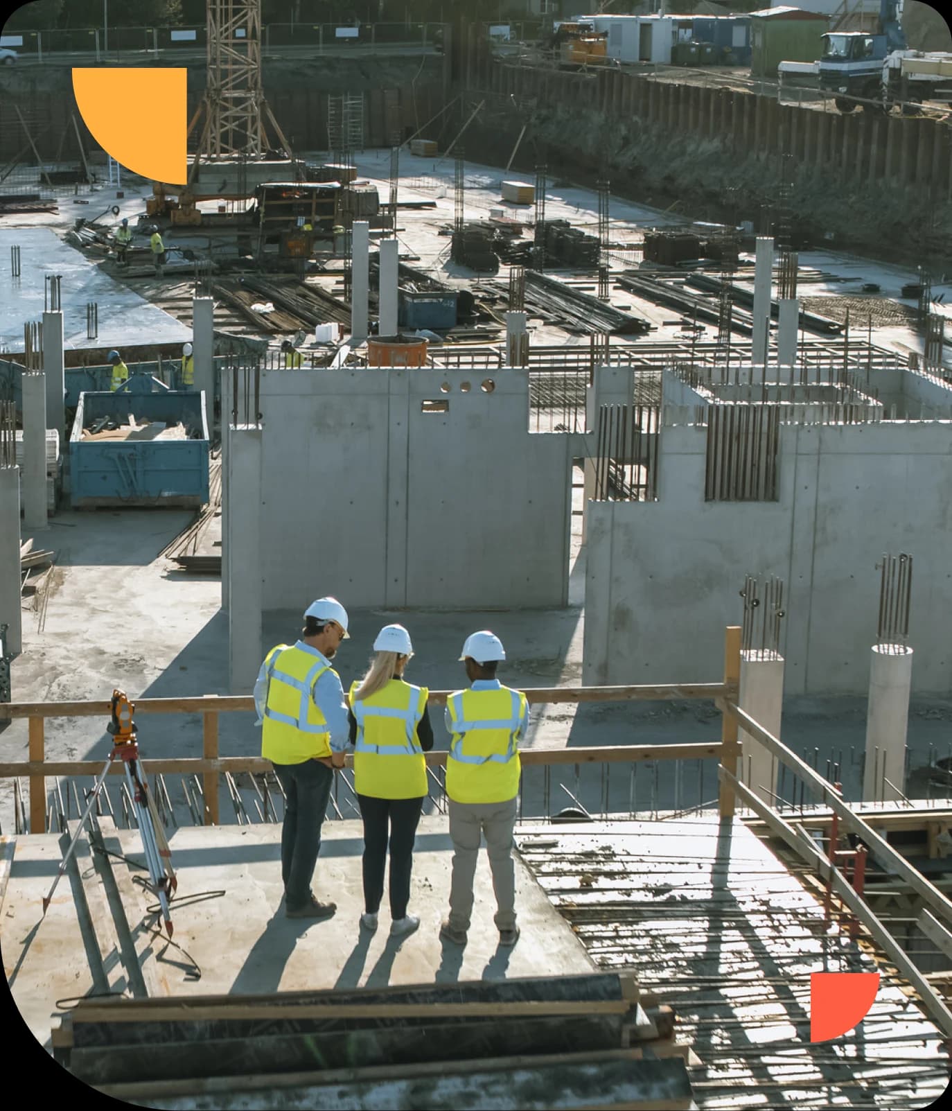 Three construction workers in safety gear and helmets stand on a platform, overlooking a large building site with concrete structures.