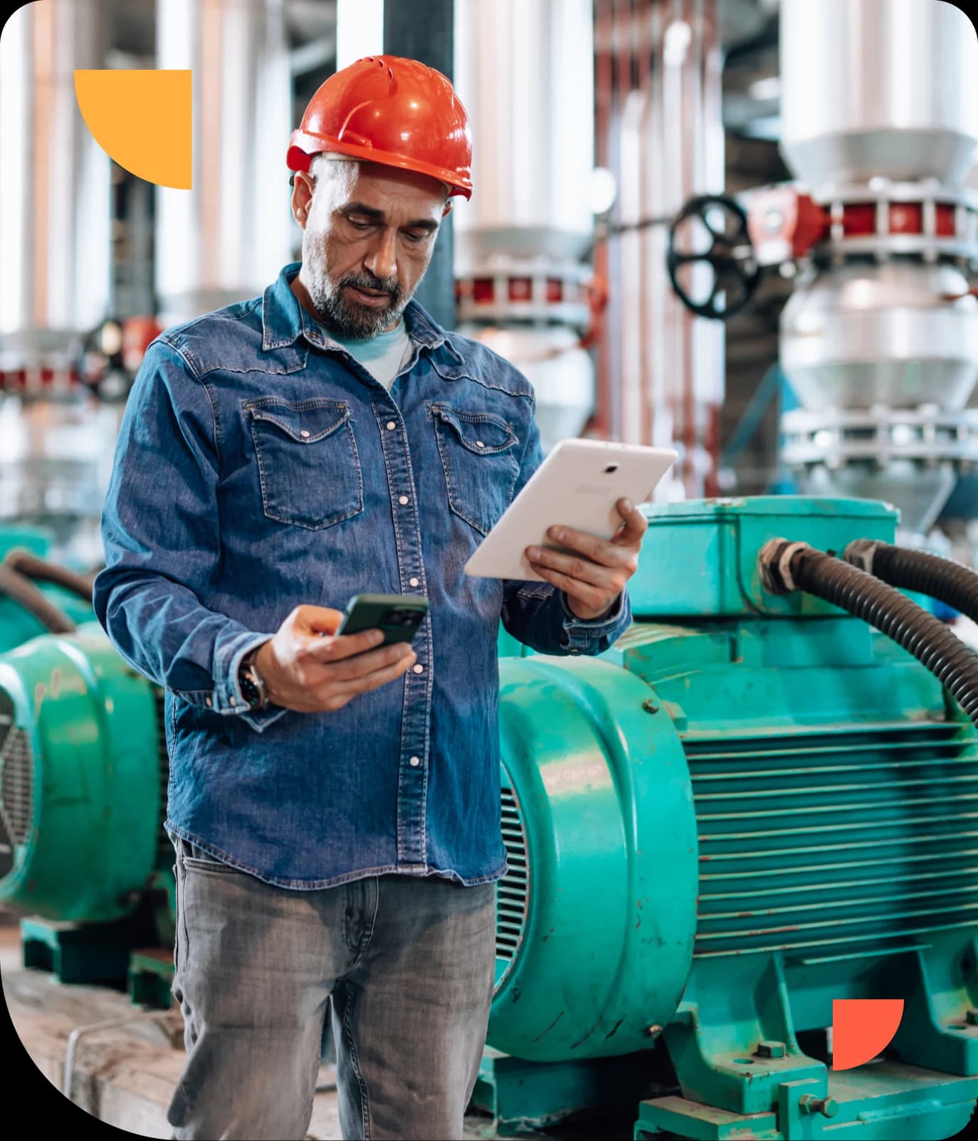 Man in a red hard hat and denim shirt using a tablet and smartphone in an industrial setting with machinery in the background.