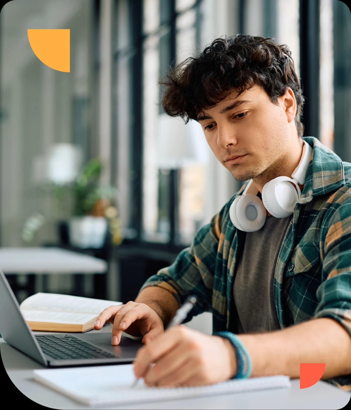 Young man with headphones around his neck, writing in a notebook, and using a laptop at a desk in a bright room.
