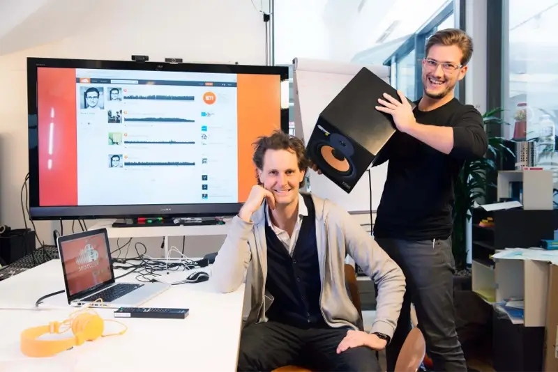 Two men in an office, one seated at a desk with a laptop and headphones, the other standing, holding a speaker. A large screen displays audio tracks.