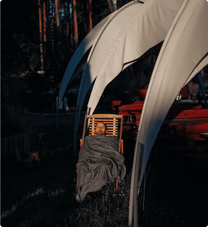 Person relaxing in a wooden chair, wrapped in a blanket, under a large white tent structure in a dark outdoor setting.
