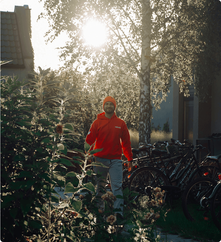 Person in a bright red hoodie and beanie walks through a garden with bicycles, backlit by the sun filtering through trees.