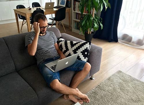 Man sitting on a gray couch using a laptop, wearing a striped shirt and shorts, with a geometric pillow beside him in a modern living room.