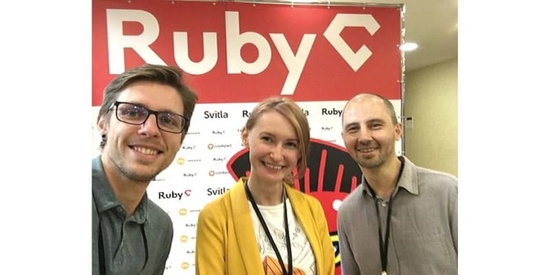 Three people smiling in front of a Ruby conference banner, featuring logos and a red and white color scheme.