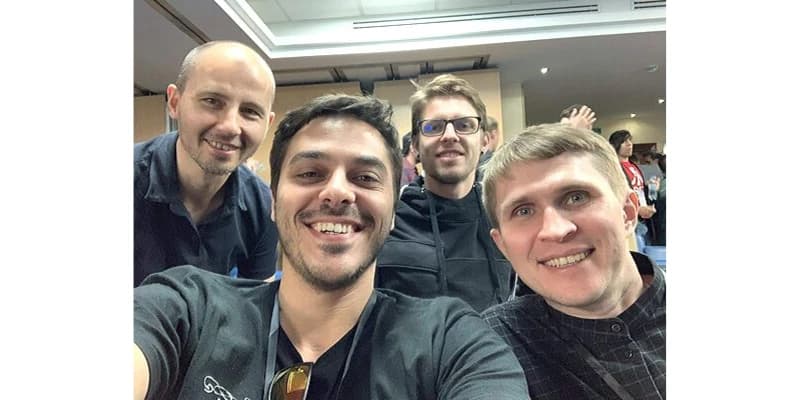 Four men smiling and posing for a selfie in a conference room, with people and a ceiling light in the background.  It's Naturaily' team.