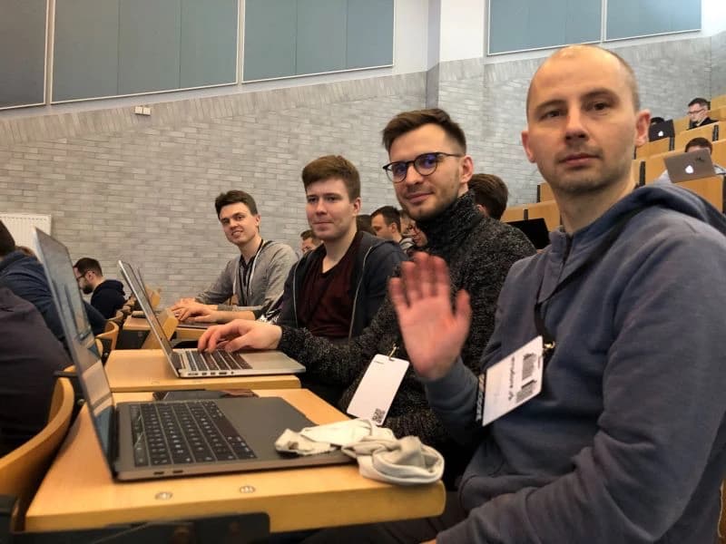 Four men sitting at desks with laptops in a lecture hall; one waves at the camera.  It's Naturaily' team on the conference.