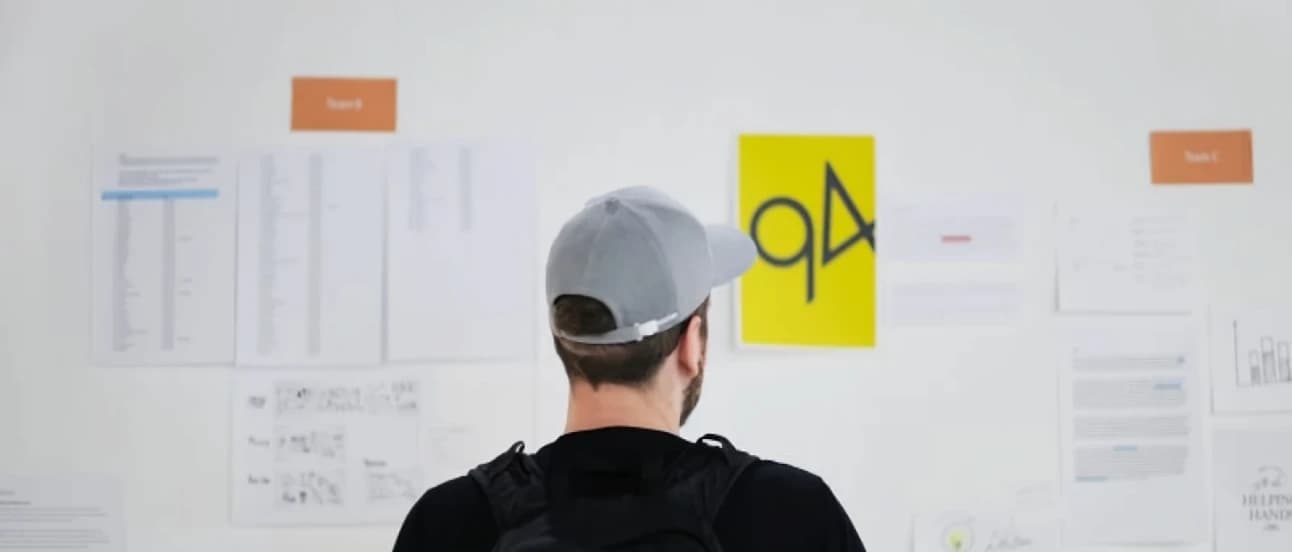 A person in a gray cap examines documents and charts on a wall, with a prominent yellow paper featuring a black symbol.